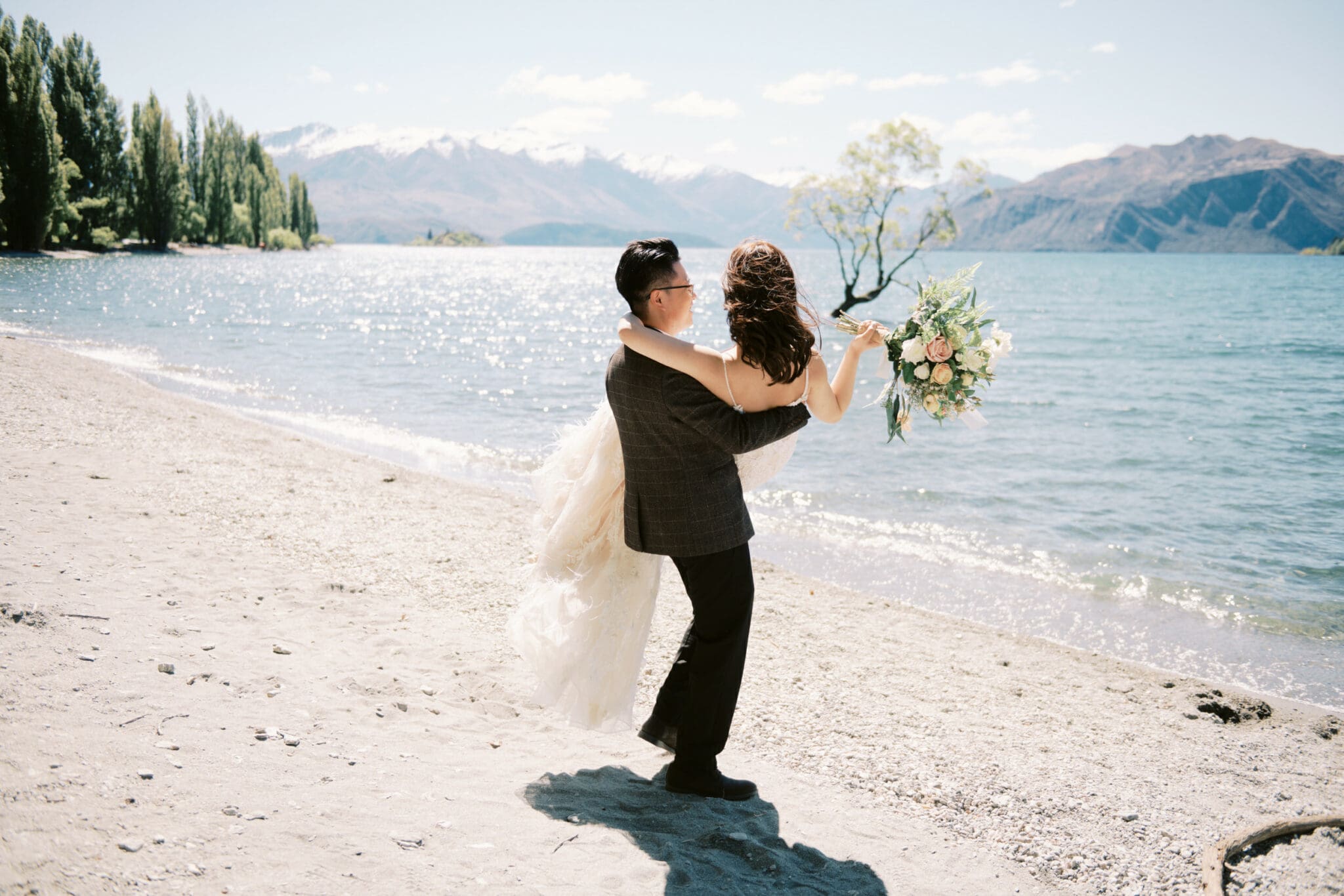 Queenstown Tekapo New Zealand Heli Wedding Elopement Pre-Wedding Shoot Photographer クイーンズタウン　テカポ　ニュージーランド　エロープメント 前撮り　フォトウェディング　結婚式 | A newlywed couple embracing on a sandy lakeshore with mountains in the background, during their pre-wedding shoot at Tekapo, New Zealand.